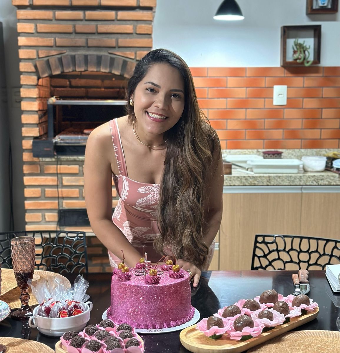 Aniversariante sorrindo diante do bolo rosa e dos docinhos preparados pela Bolateria